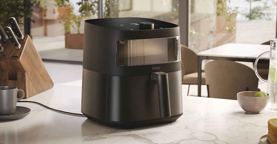 Primo piano di una friggitrice ad aria Haier I-Master Serie 5 nera su un piano di lavoro con utensili e ingredienti sullo sfondo