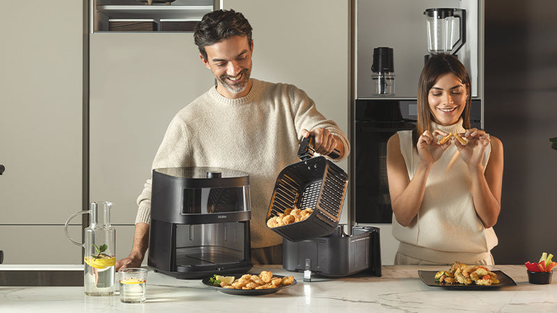 Due persone in cucina, un uomo e una donna: lui versa un ingrediente nella friggitrice ad aria Haier I-Master Serie 5 nera, lei assaggia le crocchette di pollo appena cucinate da lui. Ci sono vari alimenti sul tavolo, tra cui crocchette di pollo e verdure con pinzimonio.