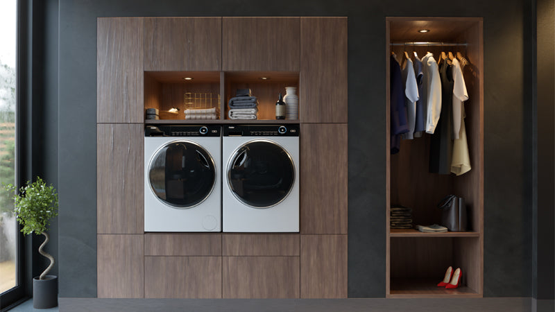 Lavanderia moderna con mobile in legno che ospita una lavatrice bianca e un’asciugatrice nera affiancate; sopra, mensole aperte con asciugamani piegati, ceste e oggetti decorativi. A destra, armadio aperto con abiti appesi, ripiani per scarpe e spazio contenitivo aggiuntivo. A sinistra, pianta in vaso sul pavimento.
