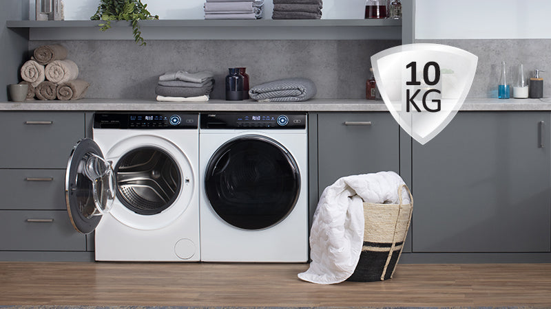 Lavanderia moderna con due lavatrici a carica frontale sotto un piano d’appoggio; una con oblò aperto, l’altra chiuso. Sopra, asciugamani piegati e decorazioni; a destra, cesto in vimini con coperta bianca e icona '10 KG'.