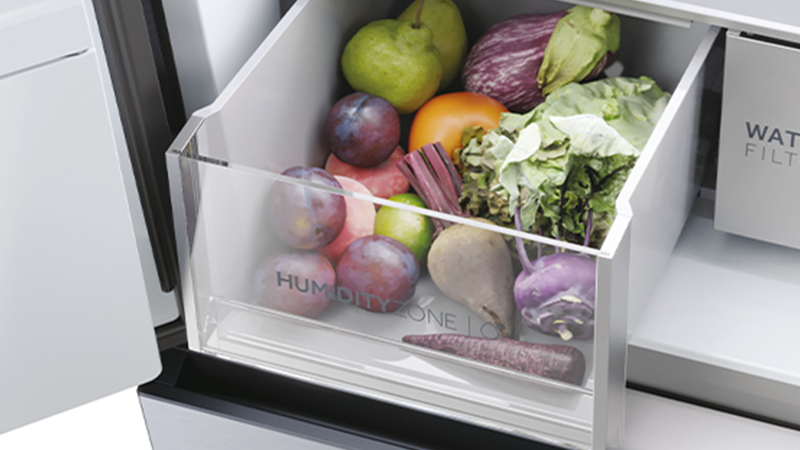 Cassetto aperto di un frigorifero etichettato HUMIDITY ZONE, contenente frutta e verdura come mele verdi, melanzane, pomodori, verdure a foglia, prugne, barbabietole e una carota; accanto, comparto con etichetta WATER FILTER.