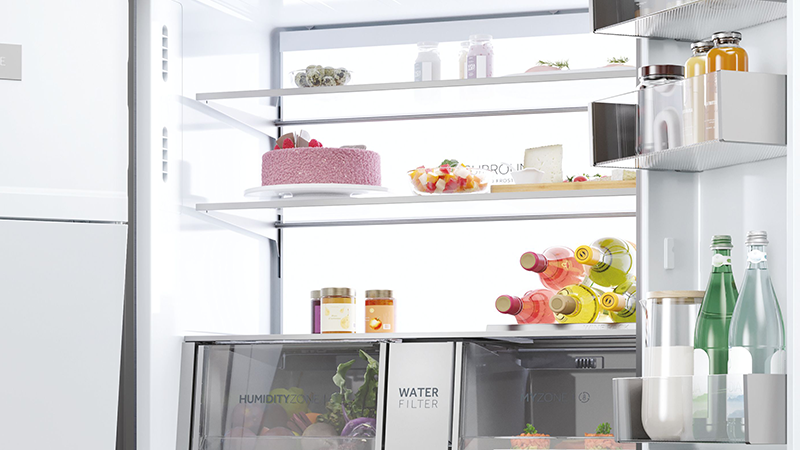 Interno di un frigorifero ordinato con torta alla fragola, insalata di frutta, conserve e bevande alcoliche e non, su diversi ripiani; comparti inferiori etichettati 'Humidity Zone' e 'Water Filter'.