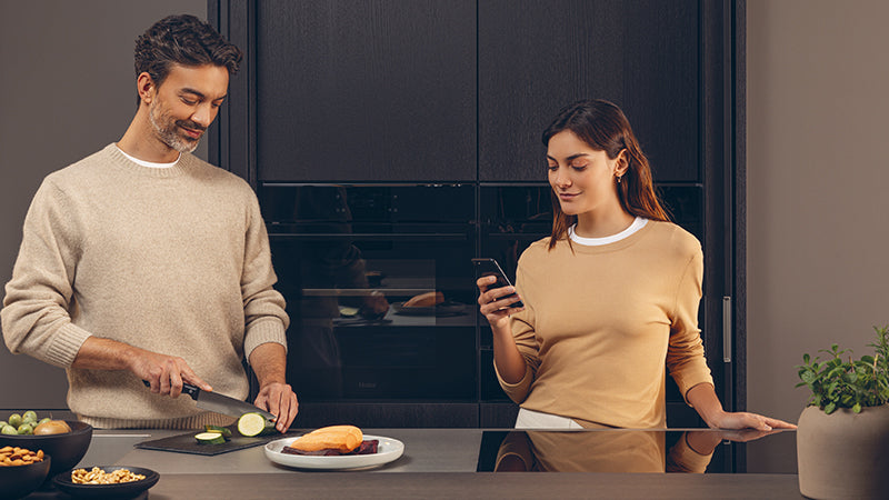 Due persone in cucina: una taglia verdure su un tagliere, l'altra guarda uno smartphone; piano di lavoro con utensili e ingredienti, sullo sfondo forno e microonde Haier