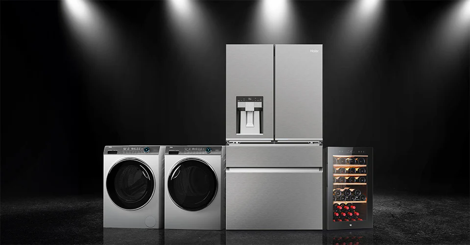 L'immagine mostra una serie di elettrodomestici moderni disposti in fila contro uno sfondo scuro, illuminati da faretti dall'alto. Da sinistra a destra si vedono: una lavatrice a carica frontale, un asciugatrice a carica frontale, un frigorifero a doppia porta (French door) con dispenser per acqua e ghiaccio integrato nella porta sinistra, e infine una cantinetta per vini con ripiani visibili contenenti bottiglie di vino. Tutti gli elettrodomestici hanno una finitura in acciaio inox lucido, conferendo un as