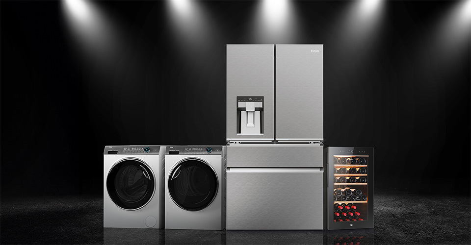 L'immagine mostra una serie di elettrodomestici moderni disposti in fila contro uno sfondo scuro, illuminati da faretti dall'alto. Da sinistra a destra si vedono: una lavatrice a carica frontale, un asciugatrice a carica frontale, un frigorifero a doppia porta (French door) con dispenser per acqua e ghiaccio integrato nella porta sinistra, e infine una cantinetta per vini con ripiani visibili contenenti bottiglie di vino. Tutti gli elettrodomestici hanno una finitura in acciaio inox lucido, conferendo un as