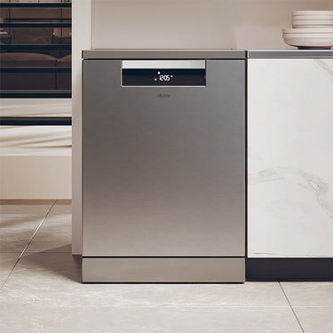 Lavastoviglie in acciaio inox integrata in una cucina moderna, con pannello di controllo digitale sul fronte e posizionata tra il frigorifero e il piano di lavoro. La cucina presenta uno stile minimalista con superfici lisce e colori neutri, e il pavimento è in piastrelle chiare.