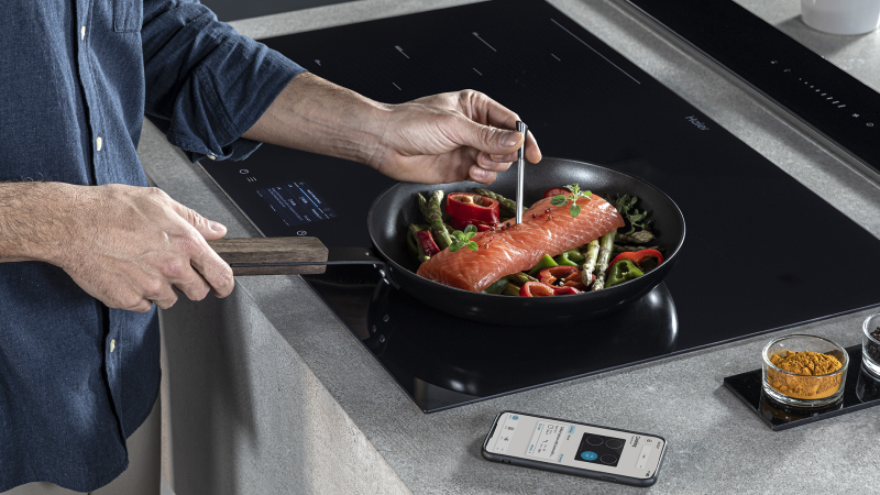 Persona che cucina su un piano a induzione ID Serie, tenendo una padella con due tranci di salmone e verdure come peperoni e fagiolini, mentre misura la temperatura con un termometro da cucina. Sul piano di lavoro, uno smartphone con un'app di cucina aperta e un contenitore di spezie.