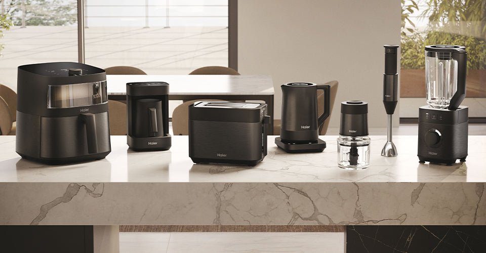 Piano cucina moderno con piccoli elettrodomestici Haier I-Master Serie 5: friggitrice ad aria, tostapane, bollitore elettrico, accessorio tritatutto e frullatore; sfondo con finestre e vista su giardino e patio