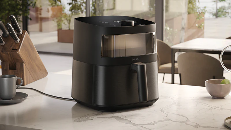 Friggitrice ad aria Haier nera con finestra trasparente su piano cucina in marmo; accanto portacoltelli in legno e ciotola con mele verdi; sfondo con tavoli e sedie vicino a grandi finestre