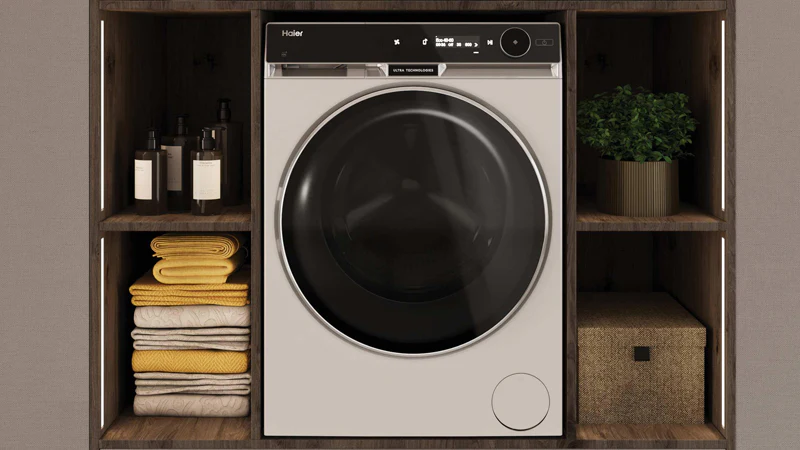 Lavasciuga Haier con oblò nero e display digitale inserita in una struttura in legno con ripiani contenenti asciugamani piegati, flaconi di detersivo, una pianta e una scatola intrecciata
