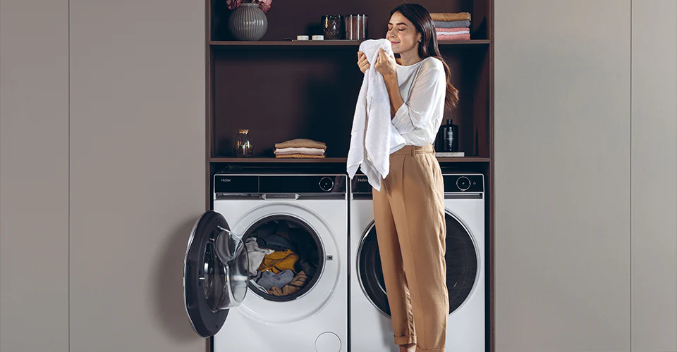 Persona davanti a una lavatrice e una asciugatrice Haier in una zona lavanderia; la lavatrice è aperta con vestiti all'interno, la persona tiene un asciugamano bianco; scaffali sopra con asciugamani piegati, contenitori e decorazioni