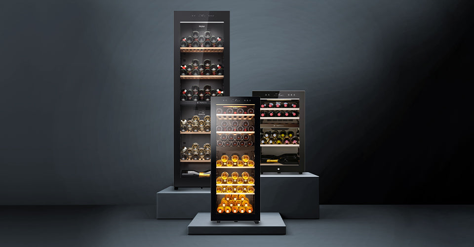 Tre modelli di cantinette vino Haier su piedistalli, con porta in vetro e bottiglie disposte su ripiani; la cantinetta centrale è illuminata, sfondo scuro che valorizza il design elegante