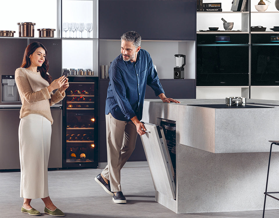 Due persone in una cucina moderna: una apre la lavastoviglie integrata nell'isola, l'altra tiene uno smartphone; sullo sfondo scaffali con utensili da cucina, cantinetta vino e forno a incasso