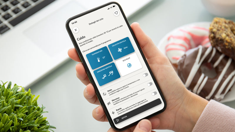 Persona tiene uno smartphone con l'app Haier aperta sulla modalità 'Caldo', visibili opzioni di climatizzazione e ventilazione