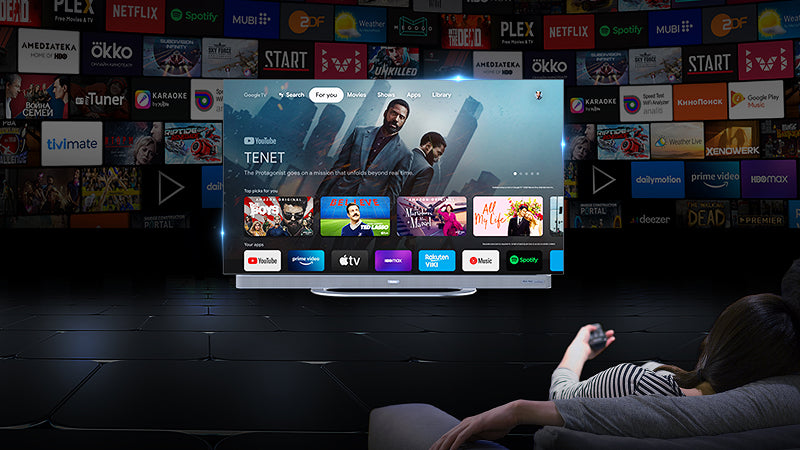 Persona seduta su un divano nel soggiorno, che utilizza un telecomando per controllare un grande schermo al centro della stanza. Il televisore mostra la schermata principale di un servizio di streaming con vari contenuti e piattaforme popolari. Sullo sfondo, una parete è piena di loghi di diverse applicazioni e servizi di streaming, creando un ambiente tecnologico e moderno.
