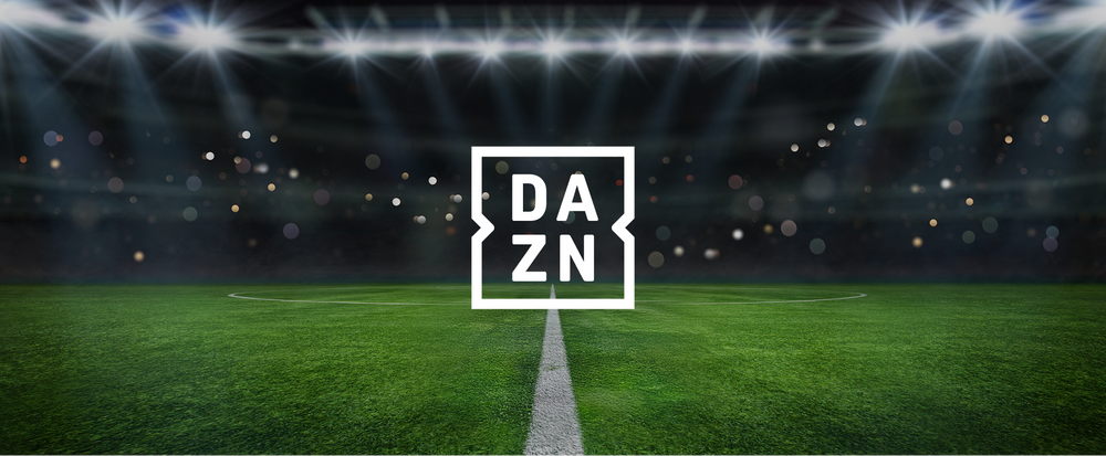 Fotografia di un campo di calcio all'aperto durante la notte, illuminato da potenti fari. Sulla superficie verde ben curata si trova il logo di DAZN al centro, con sfondo di luci soffuse e raggi di luce che si riflettono sul terreno