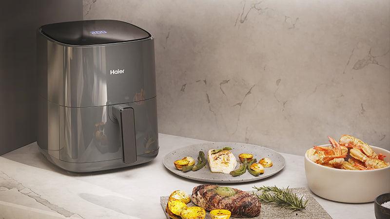 Friggitrice ad aria Haier su piano cucina con piatti di pesce grigliato, bistecca con patate e gamberi; sfondo in marmo chiaro