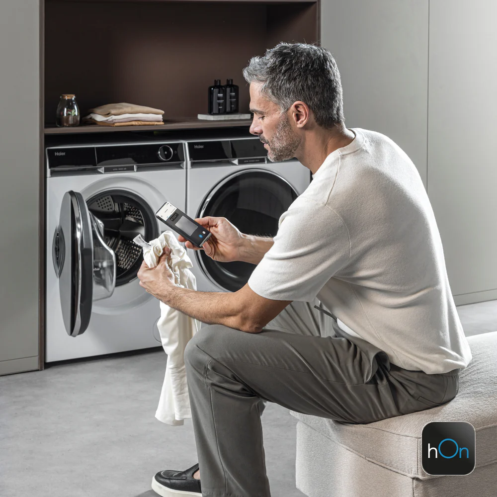 Uomo seduto su una panca imbottita davanti a due lavatrici moderne a carica frontale; tiene uno smartphone e un capo bianco, con la porta della lavatrice sinistra aperta. Sopra le macchine ci sono asciugamani piegati e flaconi. Logo 'hOn' visibile in basso a destra.