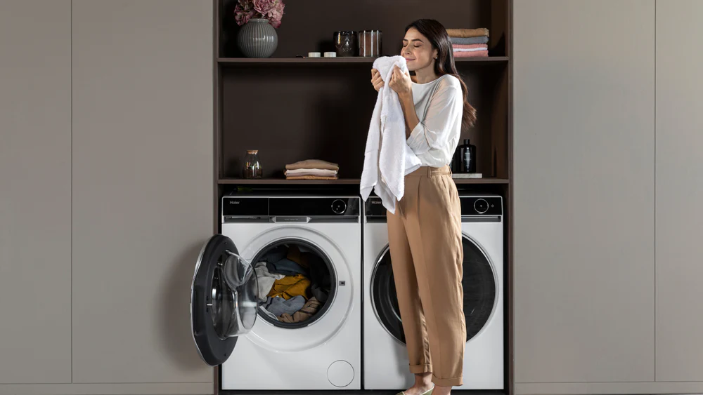 Persona davanti a lavatrice e asciugatrice moderne, impegnata nel bucato con asciugamano bianco in mano; sullo sfondo scaffalatura con asciugamani piegati, contenitori e decorazioni floreali.