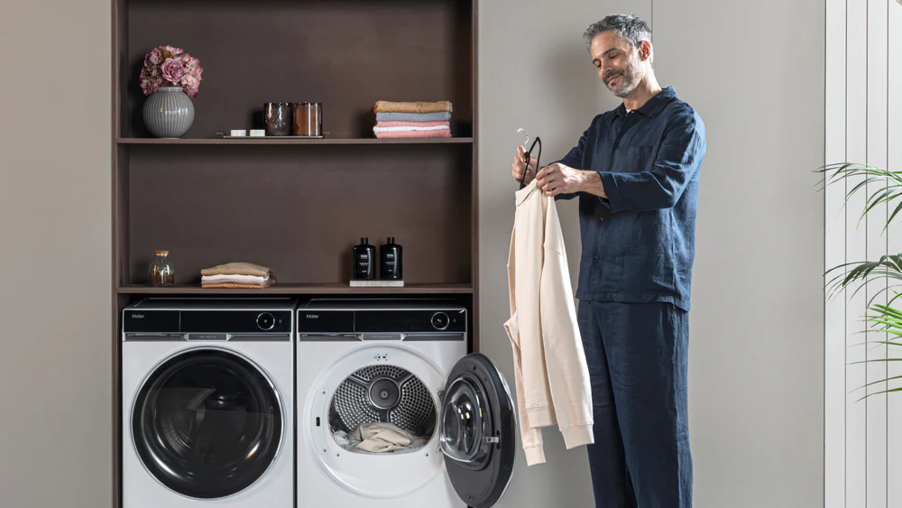 Persona accanto a lavatrice e asciugatrice moderne, mentre appende un capo su una gruccia; asciugatrice con sportello aperto e vestiti all’interno, sullo sfondo scaffale con asciugamani, contenitori e decorazioni floreali.