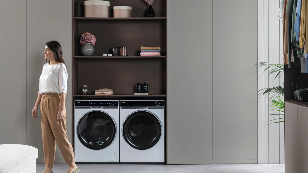 Persona accanto a lavatrice e asciugatrice moderne, con scaffali sopra contenenti asciugamani piegati, contenitori e decorazioni.