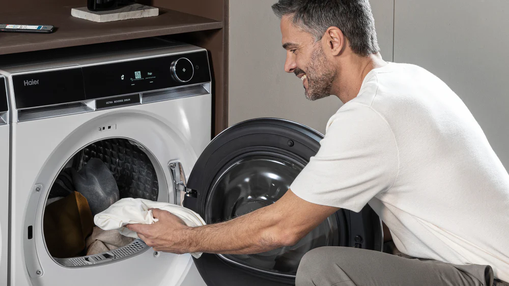 Un uomo sorridente sta caricando un bucato bianco in una lavatrice frontale moderna, di colore nero con dettagli lucidi. La lavatrice è posizionata vicino a un'altra lavatrice di colore bianco e si trova in un ambiente domestico, probabilmente una lavanderia o una stanza dedicata. L'uomo indossa una maglietta bianca e sembra felice mentre sistema i vestiti all'interno del elettrodomestico.