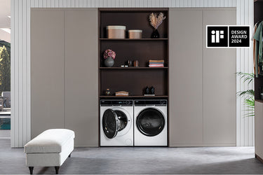 Lavanderia moderna con lavatrice e asciugatrice incassate in mobile, scaffali superiori con asciugamani piegati, vasi decorativi e contenitori; panca imbottita a sinistra, pianta a destra e logo iF Design Award 2024 in alto a destra.