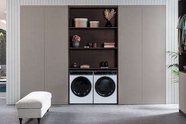 Lavanderia moderna con lavatrice e asciugatrice incassate in mobile, scaffali superiori con asciugamani piegati, vasi decorativi e contenitori; panca imbottita a sinistra, pianta a destra 