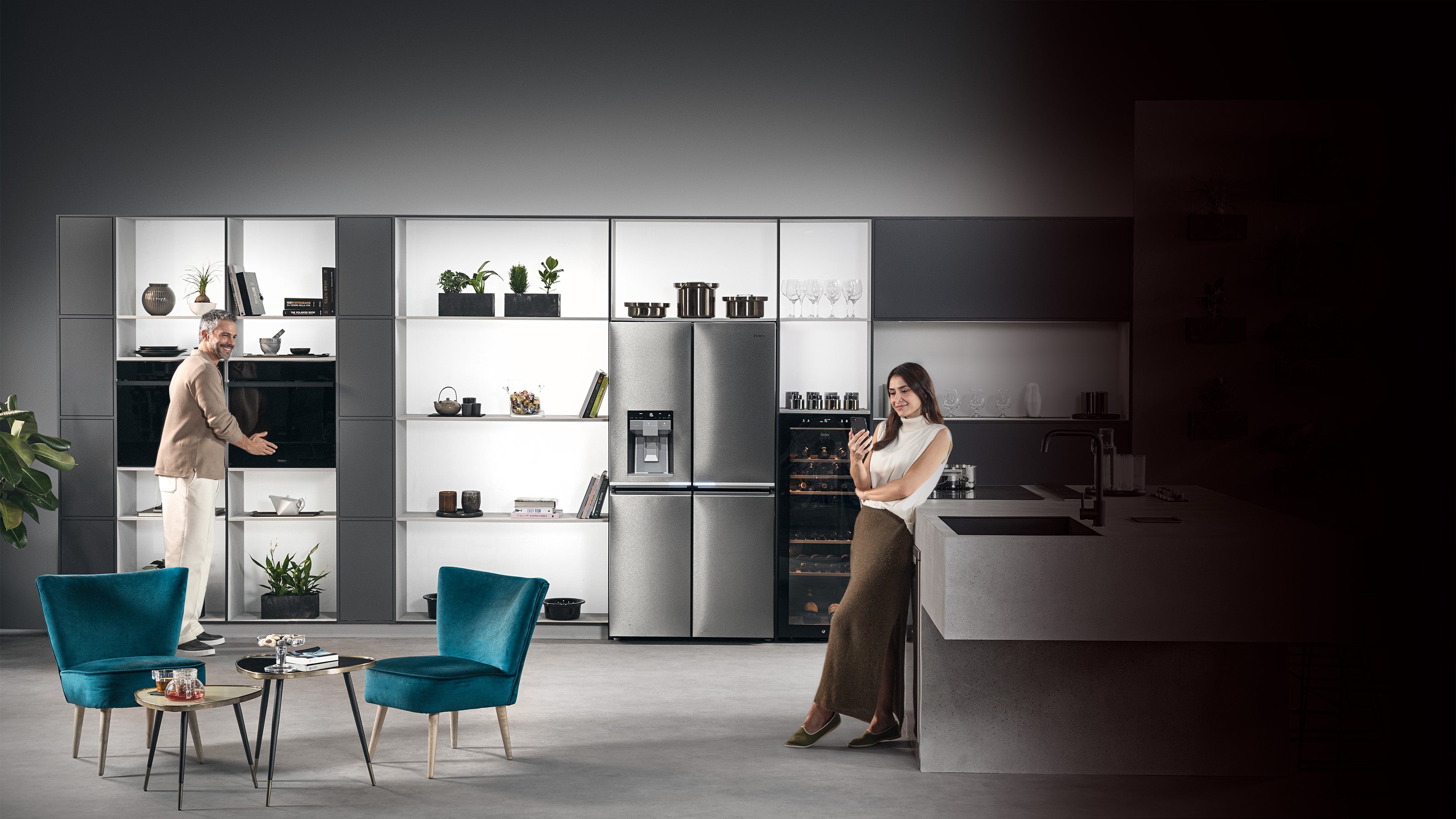 Cucina dotata di tutti gli elettrodomestici: frigorifero, microonde, forno, piano cottura a induzione. L'uomo è in prossimità del forno, la donna è appoggiata alla penisola della cucina e guarda il telefono. Tra i due ci sono due poltrone e un tavolino.