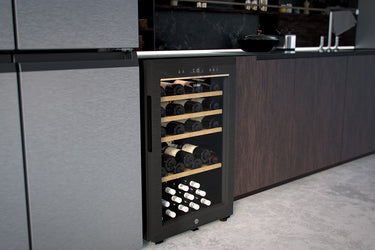 Cantinetta vino nera incassata in una cucina moderna, con porta in vetro che mostra delle bottiglie di vino posizionate su scaffali di legno. La cucina ha ante in laminato scuro e un piano di lavoro in superficie nera. Il pavimento è in cemento grigio