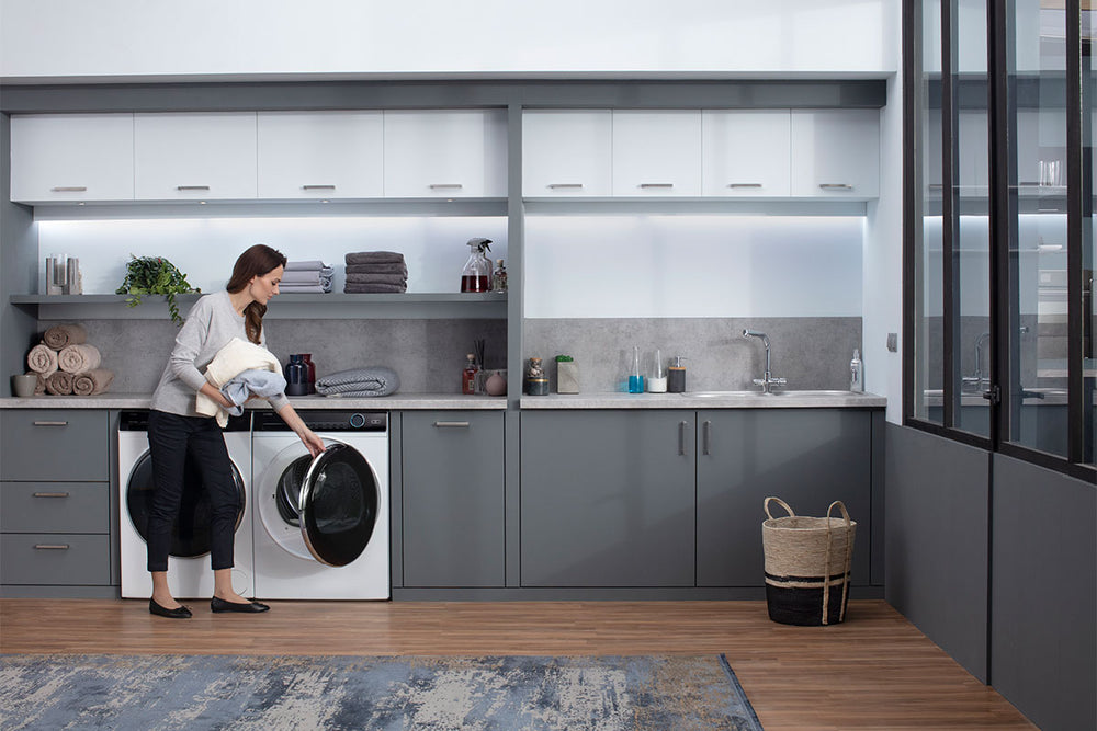 Una donna sta spostando i vestiti dalla lavatrice all'asciugatrice, disposte una di fianco all'altra. Entrambe sono inserite nella zona lavanderia di casa.