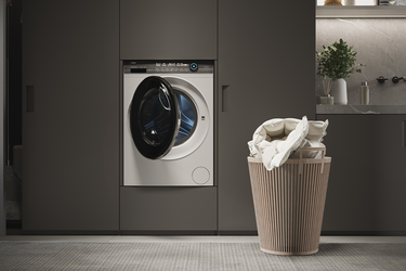 Moderna lavanderia con lavatrice a carica frontale integrata in mobili scuri, cestino della biancheria beige con vestiti bianchi in primo piano e decorazioni sul piano d'appoggio, tra cui una pianta in vaso.