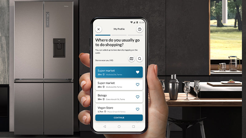 Focus su mano femminile che tiene in mano un telefono e sullo sfondo c'è un frigorifero multidoor con scompartimento del ghiaccio. Il telefono mostra l'app hon in cui si chiedono le abitudini sui negozi di acquisto. 