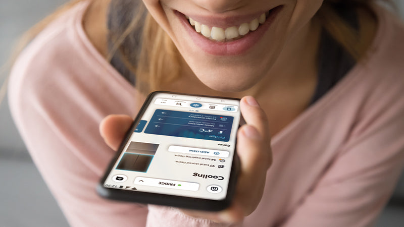 Zoom su una persona sorridente che tiene il telefono vicino alla bocca. Sul telefono si vede l'app hon e la sezione dedicata al proprio frigorifero. 