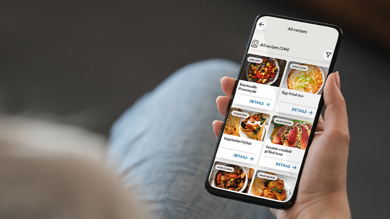 Focus su una mano che tiene in mano un telefono. Il telefono mostra le ricette presenti nell'app hon.