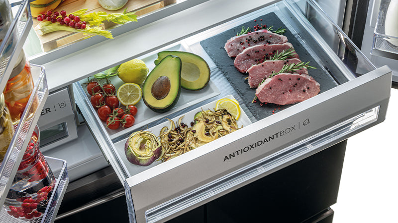 Scomparto del frigorifero "Antioxidantbox" con all'interno dei pomodori, un limone, un avocado, la carne con il pepe rosa e il rosmarino, un carciofo tagliato a fette.