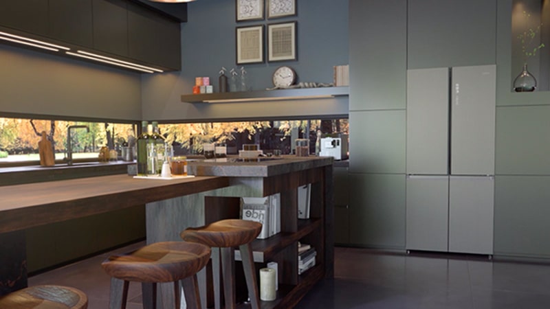 Cucina con focus sul frigorifero XXL. Di fronte al frigorifero c'è un lungo tavolo in legno con sopra una bottiglia di olio e sale. Lungo la finestra c'è il lavello e un tostapane. 