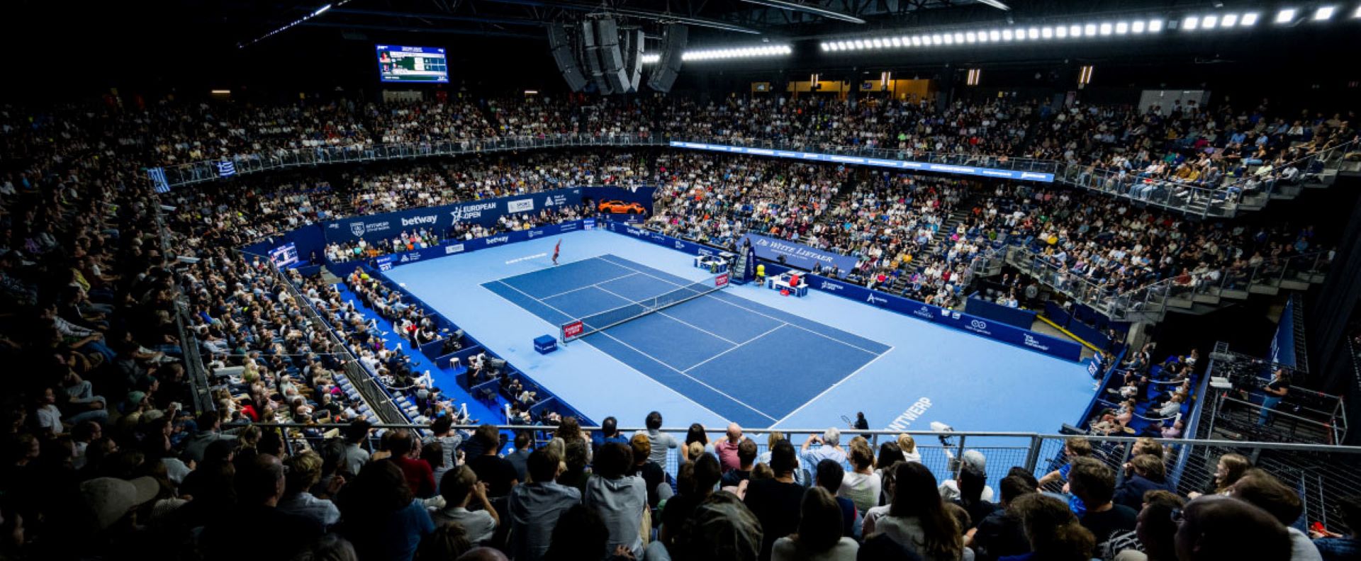 Interno di un'arena per il tennis con un campo blu e un pubblico numeroso che assiste a un match. L'illuminazione è molto intensa e l'ambiente è moderno, con schermi e strutture sopra il campo.
