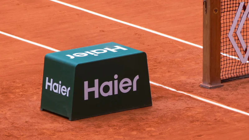 Segnaposto publicitario con la scritta 'Haier', posizionato sulla terra battuta di un campo da tennis vicino alla rete e al palo di supporto.