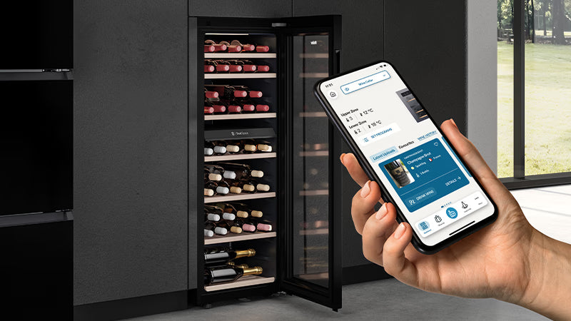 Focus su una mano femminile con in mano un telefono e sullo sfondo una cantinetta vino. Il telefono ha aperta l'app hon, in cui si può scegliere la temperatura diversificata. 